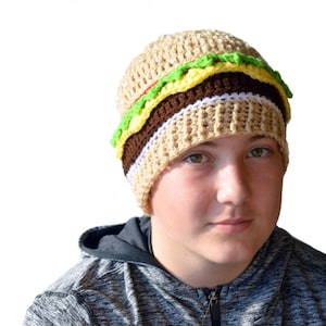 May include: A crocheted beanie hat shaped like a hamburger. The hat is brown, tan, white, green, and yellow. The hat is on a person's head.