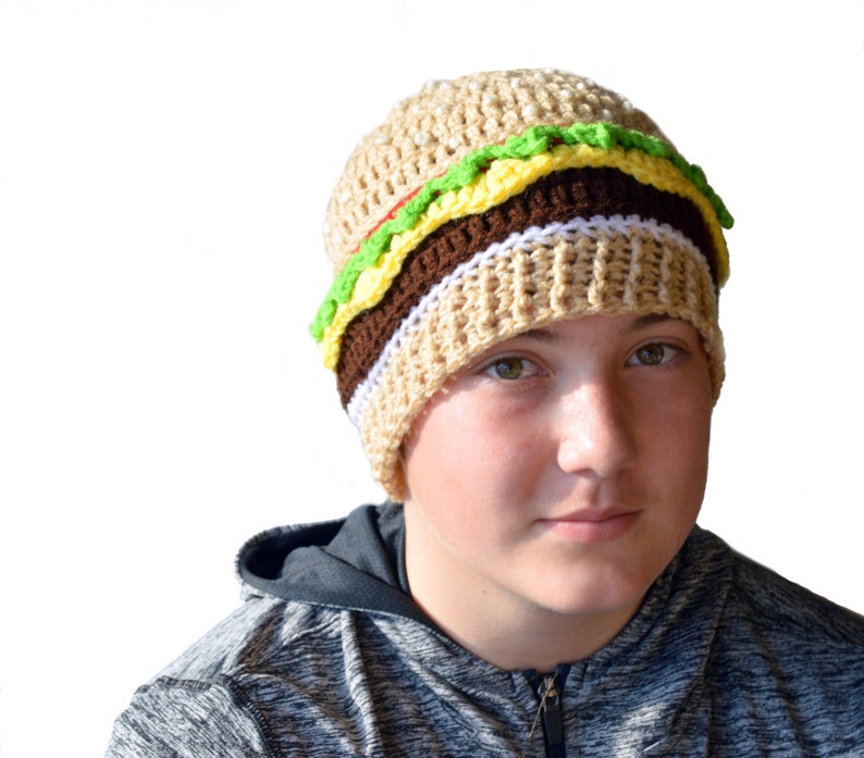 May include: A crocheted beanie hat shaped like a hamburger. The hat is brown, tan, white, green, and yellow. The hat is on a person's head.