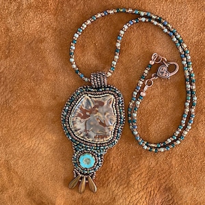 May include: A beaded necklace with a wolf pendant. The pendant is made of brown and white ceramic with a turquoise stone accent. The necklace is made of copper and green beads.