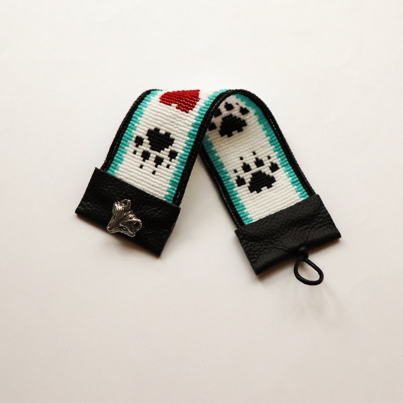 May include: A white and turquoise beaded strap with black paw prints and a red heart. The strap has black leather ends with a silver paw print charm.