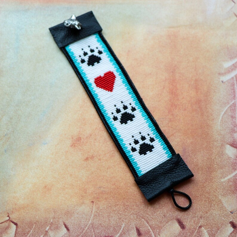 May include: White beaded bracelet with black bear paw prints and a red heart. The bracelet has a black leather closure with a silver charm.