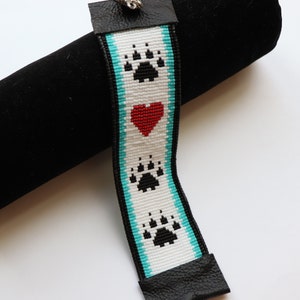 May include: A white beaded bracelet with a turquoise border and three black paw prints. A red heart is centred between the top and bottom paw prints.