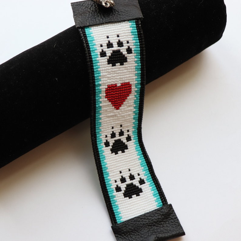 May include: A white beaded bracelet with a turquoise border and three black paw prints. A red heart is centred between the top and bottom paw prints.