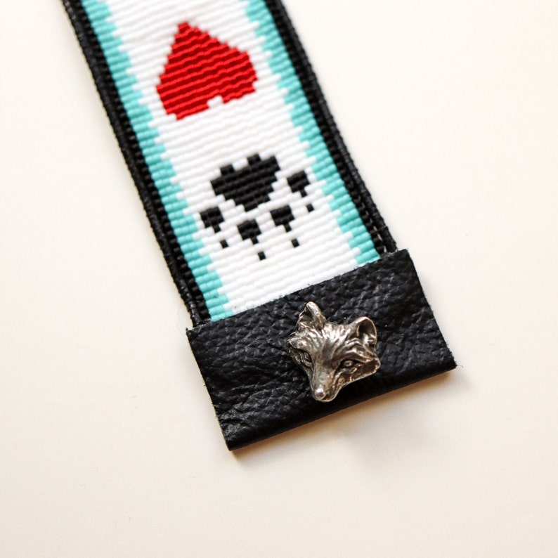 May include: A beaded strap with a red heart, black paw print, and turquoise accents. The strap is attached to a black leather tab with a silver fox head charm.