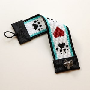 May include: A white beaded bookmark with black paw prints, a red heart, and a silver fox charm. The bookmark is attached to black leather tabs at each end.