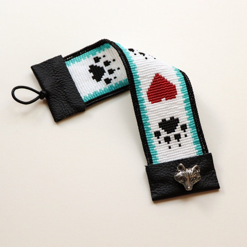 May include: A white beaded bookmark with black paw prints, a red heart, and a silver fox charm. The bookmark is attached to black leather tabs at each end.