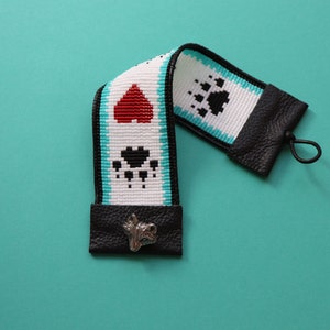 May include: A white and black beaded camera strap with red heart and paw print designs. The strap has black leather ends with a silver charm.