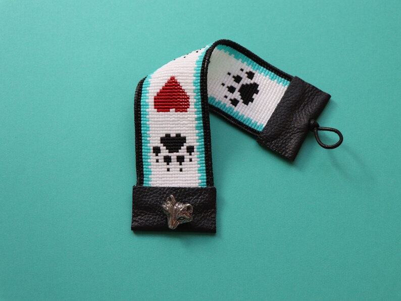 May include: A white and black beaded camera strap with red heart and paw print designs. The strap has black leather ends with a silver charm.