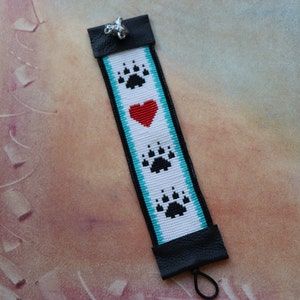 May include: A white beaded bracelet with a turquoise border and black bear paw prints. A red heart is centred between the paw prints. The bracelet has a black leather closure with a silver charm.