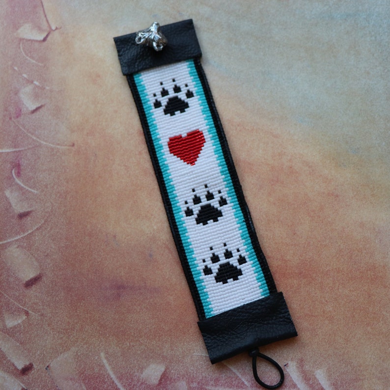 May include: A white beaded bracelet with a turquoise border and black bear paw prints. A red heart is centred between the paw prints. The bracelet has a black leather closure with a silver charm.