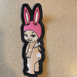 May include: An embroidered patch featuring a cartoon character with tan skin, wearing a bright pink bunny ear hat. The character has small white wings on its back and white spots on its body, all outlined in a thick black border with stitching.