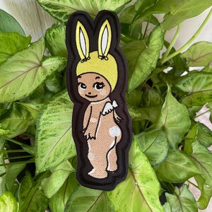 May include: An embroidered patch featuring a cartoon character with tan skin, wearing a yellow bunny ear hat. The character has small white wings on its back, a white tail, and is outlined in a thick black border against a dark background.