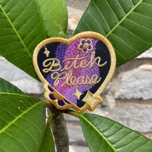 May include: A heart-shaped embroidered patch with a gold border. The background features a gradient of purple and black threads. The text "Bitch Please" is stitched in gold cursive script, accompanied by small gold star and flower motifs.