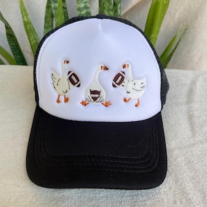 Puede incluir: Una gorra tipo trucker negra y blanca con un diseño bordado de tres gansos blancos con patas naranjas, cada uno sosteniendo un balón de fútbol americano marrón. La gorra tiene una visera negra, un panel frontal blanco y malla negra trasera.