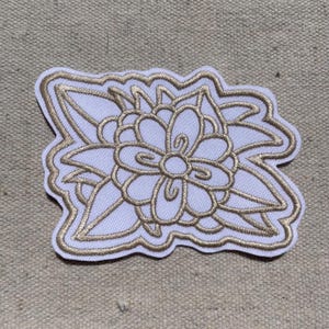 May include: An embroidered patch featuring a white fabric base with a central floral design outlined in metallic gold thread. The flower has rounded petals and is surrounded by pointed leaves, all defined by thick, raised gold embroidery stitching.