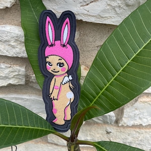 May include: An embroidered patch featuring a cartoon character wearing a pink bunny ear hat. The character has a tan body, pink spots, and small white wings on its back. The design is outlined in a thick black border with a dark navy blue edge.
