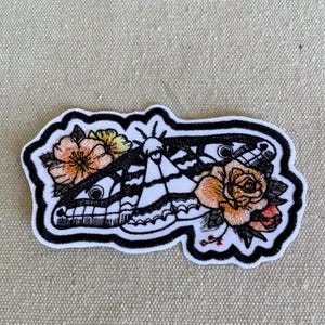 May include: An embroidered patch featuring a black and white moth with detailed wing patterns, flanked by peach and yellow flowers with green leaves. The design is outlined in a thick black border on a white background, creating a high-contrast look.