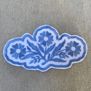 May include: An embroidered patch featuring a white background with a scalloped edge and a central floral design. The motif consists of three blue flowers with stems and leaves, all rendered in blue thread against the white fabric base.
