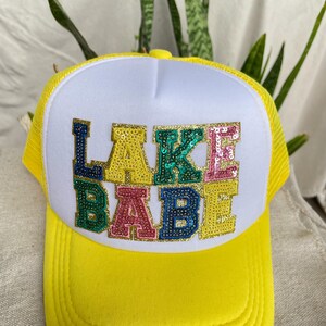 May include: A bright yellow trucker hat with a white front panel featuring the text LAKE BABE in large, multi-colored sequined letters. Each letter is outlined in gold glitter, with the sequins appearing in blue, yellow, green, and pink.