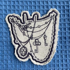 May include: An off-white fabric patch featuring dark purple embroidered outlines and details. The design depicts a series of draped chains, a circular pendant with an intricate center, and two hanging keys, all rendered in a dark purple thread.