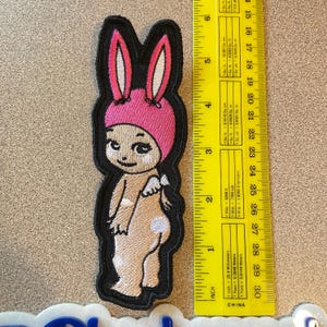 May include: Embroidered patch featuring a cartoon baby wearing a pink bunny hat with long ears. The baby has small white wings and a white spot on its bottom. The patch is outlined in black and is approximately 4 inches tall.