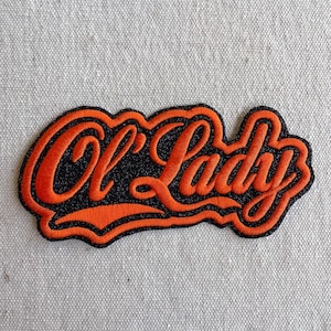 May include: An embroidered patch featuring the text Ol' Lady in a cursive script. The lettering is bright orange with a thick orange border, set against a black glittery background. The patch has a distinct, irregular shape that follows the text.