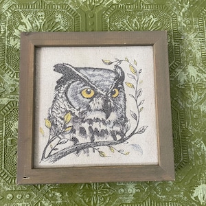 May include: Square framed artwork featuring a detailed illustration of an owl with yellow eyes, gray and white feathers, and a branch with yellow leaves. The artwork is set against a neutral linen background and framed in a gray wooden frame.