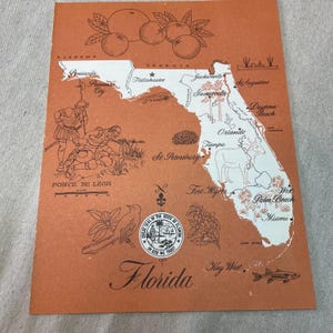 May include: Vintage Florida map illustration on orange paper. Features oranges, state seal, and drawings of Ponce de Leon and a fish. Includes city names like Miami and Jacksonville.