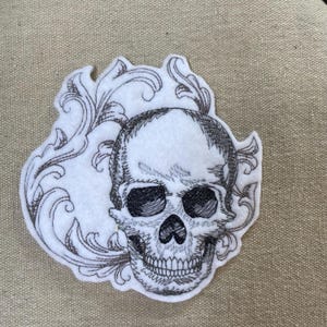 May include: A white fabric patch featuring a black embroidered skull design surrounded by intricate, swirling filigree patterns. The embroidery uses fine lines to create shading and texture on the skull, set against a plain white felt-like background.