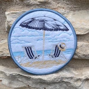 May include: Embroidered round patch featuring a beach scene. The design includes a beach umbrella, two beach chairs, and a hat. The background depicts a blue sky and ocean. The patch is bordered with a blue stitched edge.