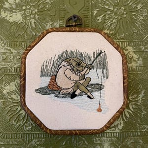 May include: Embroidered artwork in an octagonal wooden frame. The artwork depicts a frog-like creature fishing, wearing a coat and seated on a small woven mat. The background features tall grass and a fishing line with a bobber.