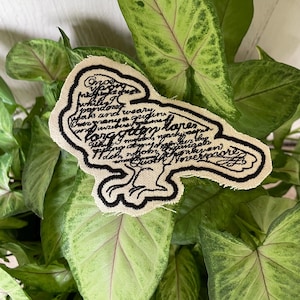 May include: A beige fabric patch shaped like a raven, featuring black embroidered outlines and black cursive text from Edgar Allan Poe's The Raven, including the phrase Quoth the Raven Nevermore, with a thick black border and frayed edges.