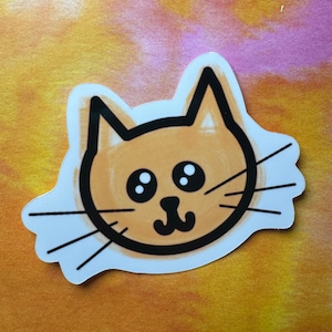May include: A cartoon cat sticker with a light orange face, black eyes, and a black nose. The cat has a black outline and white whiskers.