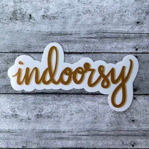 Op de afbeelding: Een witte sticker met een gouden omtrek van het woord "indoorsy" geschreven in een cursief schrift.