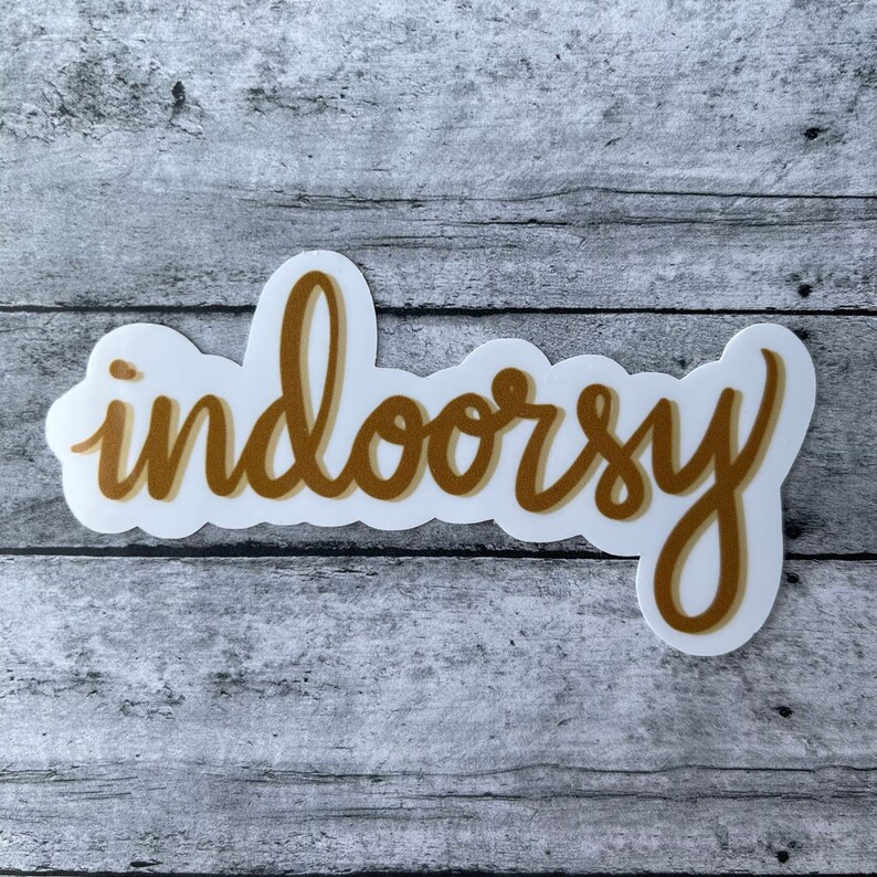 Op de afbeelding: Een witte sticker met een gouden omtrek van het woord "indoorsy" geschreven in een cursief schrift.