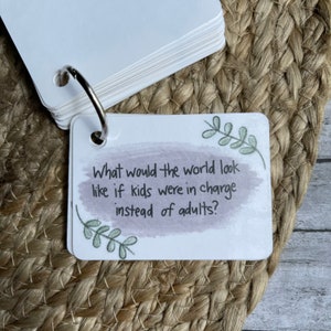 May include: A white card with a purple watercolor background and green leaves on either side. The card has the text "What would the world look like if kids were in charge instead of adults?"