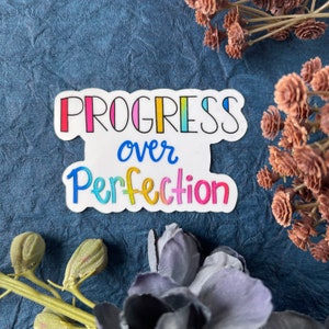 May include: A white sticker with the text "Progress over Perfection" in rainbow colors. The sticker is on a blue textured background.