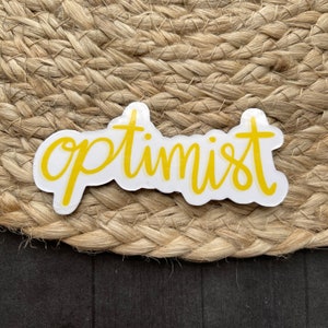 May include: Yellow vinyl sticker with the word "optimist" written in a cursive font.