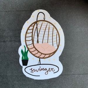 May include: A white sticker with a brown wicker hanging chair with a pink cushion. The chair is surrounded by yellow lights and the word "swinger" is written in cursive below the chair.