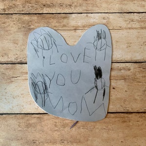 May include: A handmade gray paper card with the words "I LOVE YOU MOM" written in black marker. The card has handprints drawn around the text. The card is on a wooden surface.