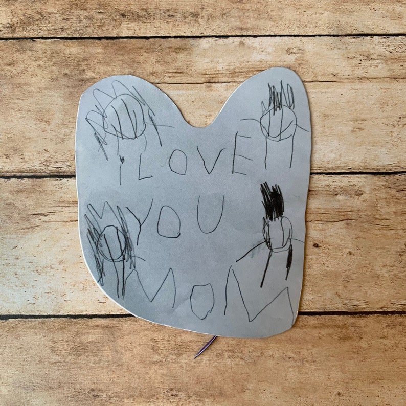 May include: A handmade gray paper card with the words "I LOVE YOU MOM" written in black marker. The card has handprints drawn around the text. The card is on a wooden surface.
