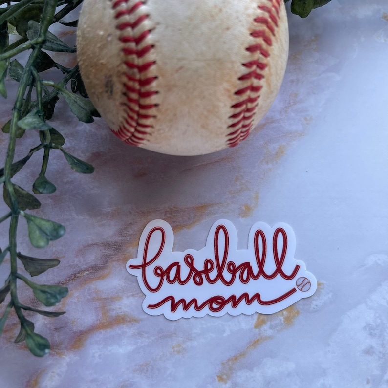 May include: A white sticker with red cursive text that says "baseball mom" with a small baseball graphic next to the text. A baseball is in the background.