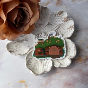 May include: A brown and green house with a rounded door and a green roof covered in mushrooms. The house has a chimney with smoke coming out of it. The house is surrounded by green grass and a fence.