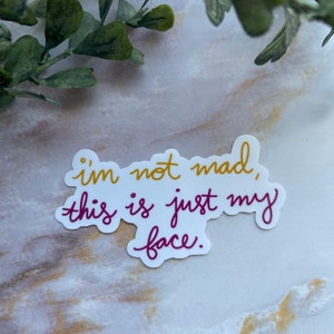 May include: White sticker with the text "i'm not mad, this is just my face." in gold and purple cursive lettering.