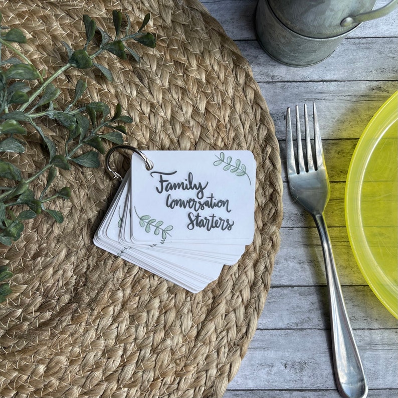 May include: A set of white cards with green leaf illustrations and the text "Family Conversation Starters" on the top card. The cards are fanned out and held together with a silver ring.