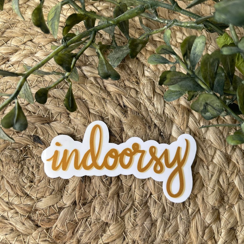 Op de afbeelding: Een witte sticker met het woord "indoorsy" geschreven in een geelgouden cursief schrift.