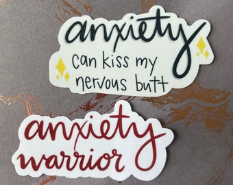 May include: Two white stickers with black and red text. One sticker says "anxiety can kiss my nervous butt" with yellow star accents. The other sticker says "anxiety warrior" in a cursive font.