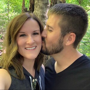 以下が含まれることがあります： 森の中に立っているカップル。男性が女性の頬にキスをしている。女性はグレーのタンクトップを着て、男性は黒いTシャツを着ている。