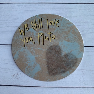 May include: A round sticker with a blue and brown marbled background. The sticker has a heart shape in the center and the text "We still love you, Pluto." in yellow lettering.