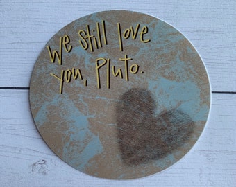 Op de afbeelding: Een ronde sticker met een textuur van lichtblauw en beige met een donkerbruine hartvorm rechtsonder. De tekst "we still love you, Pluto." is in een donker, handgeschreven lettertype in het bovenste gedeelte gedrukt.